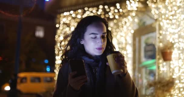 Schöne modische glücklich stilvolle junge Frau in braunem Mantel mit dunkelblauem Schal und langen dunkelgelockten Haaren genießt Kaffee beschäftigt mit ihrem Mobiltelefon, während sie eine Straße in der Stadt spaziert. hübsche Mädchen-Nachricht — Stockvideo