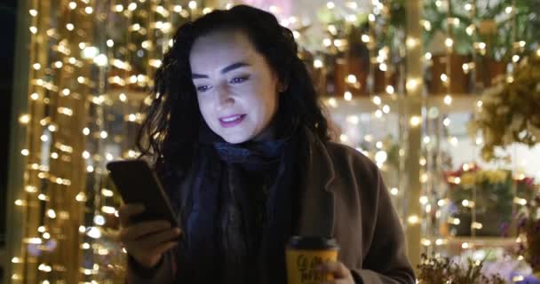 Belle jeune femme élégante heureuse et à la mode en manteau marron avec écharpe bleu foncé et longs cheveux bouclés foncés apprécie le café occupé avec son téléphone portable tout en marchant dans une rue de la ville. Jolie fille Messagerie — Video