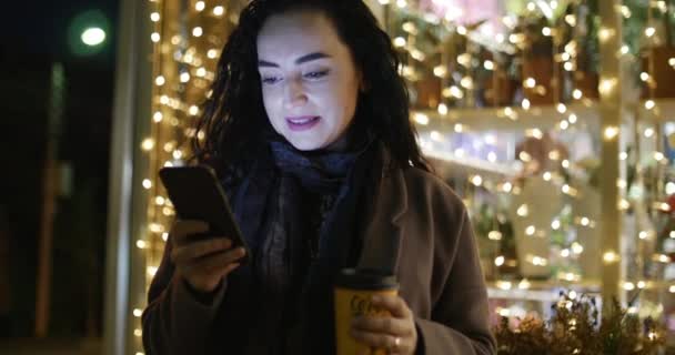 Krásná módní Happy elegantní mladá žena v Brown kabát s tmavě modrý šátek a dlouhé tmavé kudrnaté vlasy má káva zaneprázdněn s její mobilní telefon při chůzi ulice města. Hezká dívka Messaging — Stock video