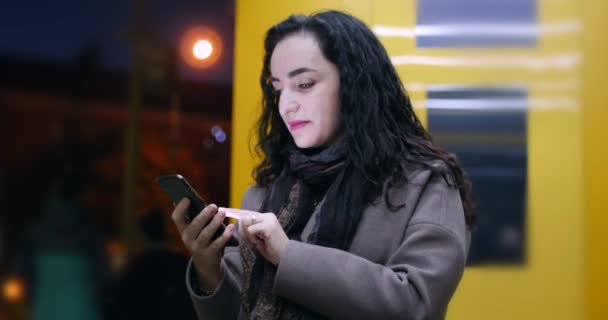 Schöne glückliche junge hübsche Frau in braunem Mantel mit dunkelblauem Schal und langen dunklen Haaren genießt Kaffee-Nachrichten auf dem Smartphone auf dem Hintergrund der Nacht oder Abend Stadt. — Stockvideo