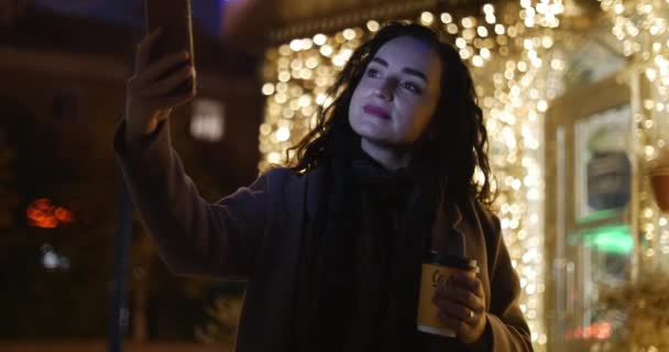 Hermosa joven feliz Linda chica de abrigo marrón con bufanda azul oscuro y largo pelo rizado oscuro disfruta del café Ocupado con su teléfono móvil Mujer hace una selfie en el fondo de las luces festivas de — Vídeos de Stock