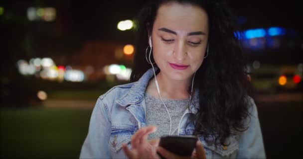 Счастливая стильная молодая симпатичная девушка в белой куртке с белыми наушниками и длинным темным хайром на смартфоне, слушающая музыку на фоне ночи или развивающегося города — стоковое видео
