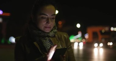 Güzel mutlu şık genç güzellik bir koyu mavi eşarp ve uzun siyah saçlı kahverengi deri ceketli bir gece veya akşam şehir çerçevede Smartphone iletilerde sahiptir. 4k.