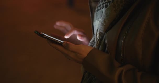 Belle belle jeune beauté élégante en cuir marron veste avec une écharpe bleu foncé et cheveux longs foncés bénéficie de messages sur le Smartphone sur le fond d'une nuit ou d'une ville du soir. 4K . — Video