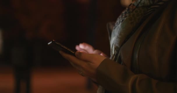 Belle belle jeune beauté élégante en cuir marron veste avec une écharpe bleu foncé et cheveux longs foncés bénéficie de messages sur le Smartphone sur le fond d'une nuit ou d'une ville du soir. 4K . — Video