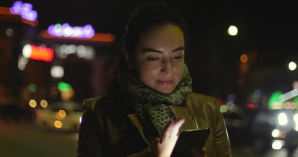 Bela beleza jovem e elegante feliz em jaqueta de couro marrom com um cachecol azul escuro e cabelo escuro longo goza de mensagens no smartphone contra o fundo de uma noite ou cidade à noite. 4K . — Vídeo de Stock