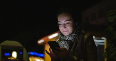 Güzel mutlu şık genç güzellik kadın kahverengi deri ceketli bir koyu mavi eşarp ve uzun siyah saçlı bir gece veya akşam şehir çerçevede Smartphone iletilerde sahiptir. 4k