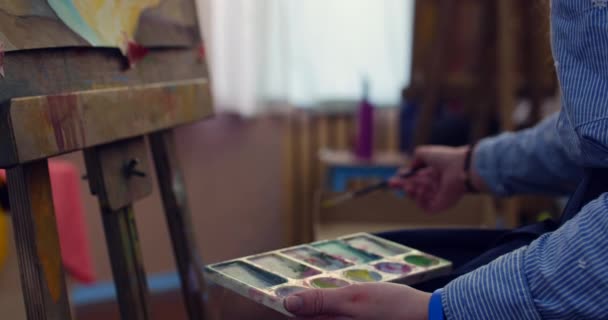 Una joven y hermosa artista femenina está en un estudio de arte, sentada detrás de un caballete y pintando sobre lienzo. Proceso de Dibujo: en el Estudio de Arte de la Mano de los Artistas Chica de Arte con Pincelada sobre Lienzo.4K — Vídeos de Stock