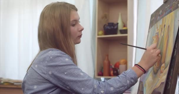 Een jonge mooie vrouwelijke kunstenaar ligt in een Art Studio, zitten achter een ezel en schilderen op Canvas. Tekening proces: in het Art-Atelier van de kunstenaars Hand kunst meisje met een borstel schilderij op Canvas.4k — Stockvideo