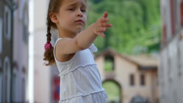 A sweet little curly girl with golden hair is sad and at the same time spinning in the dance and rotating in the street against the backdrop of the mountains. Average deceleration. — Stock Video