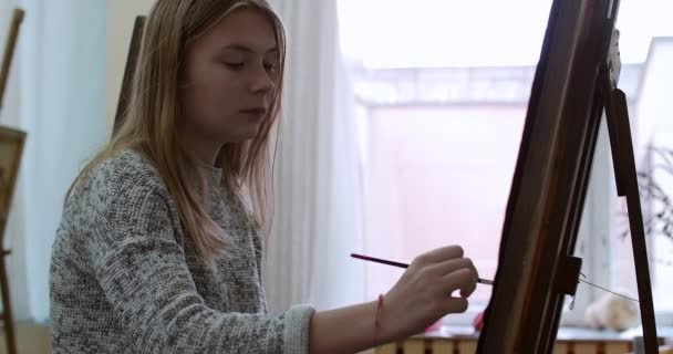 Jonge mooie vrouwelijke artiest is een Art Studio, zitten achter een ezel en schilderen op Canvas. Tekening proces: in het Art-Atelier van de kunstenaars Hand kunst meisje met een borstel schilderij op Canvas.4k — Stockvideo