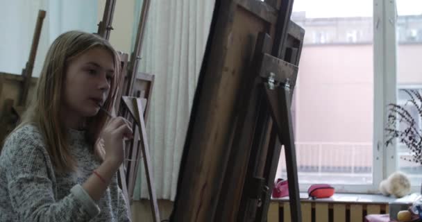 Die junge schöne Künstlerin sitzt in einem Kunstatelier hinter einer Staffelei und malt auf Leinwand. Zeichenprozess: im Atelier des Künstlers Hand Art Girl mit einem Pinsel Malerei auf Leinwand. 4k — Stockvideo