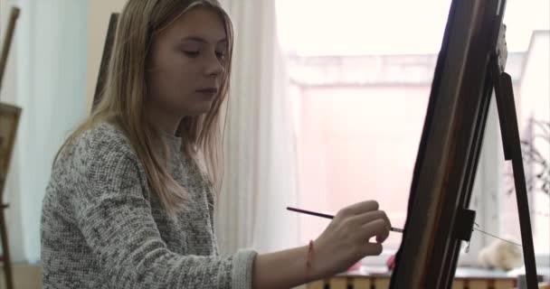 Jonge mooie vrouwelijke artiest is een Art Studio, zitten achter een ezel en schilderen op Canvas. Tekening proces: in het Art-Atelier van de kunstenaars Hand kunst meisje met een borstel schilderij op Canvas.4k — Stockvideo