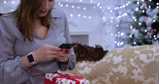 Νεαρή γυναίκα όμορφη βραδιά των Χριστουγέννων γράφοντας μηνύματα κειμένου από κινητό τηλέφωνο, κάθεται στον καναπέ στο σπίτι, με φόντο τα φώτα των Χριστουγέννων. — Αρχείο Βίντεο