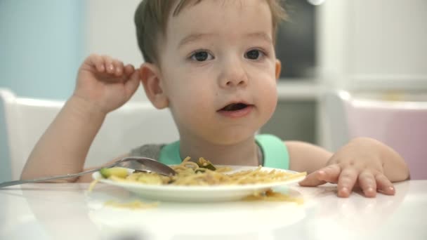 Małe dziecko siedzi przy stole w szelkach i jeść jego własne spaghetti, dziecko chętnie zjada. — Wideo stockowe