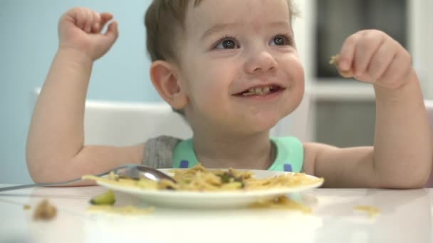Criança pequena está sentada a uma mesa em um babador e comer seu próprio espaguete, o bebê come de bom grado. Instrução . — Vídeo de Stock