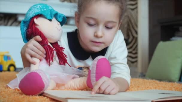 Menina bonito com cabelo encaracolado, em pijamas infantis, deitado no chão em um tapete de malha e ela mesma lendo um livro infantil. Menina lendo um livro abraçando sua boneca bebê macio . — Vídeo de Stock