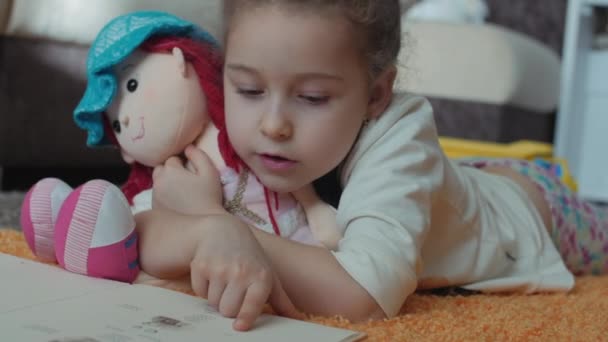Söt liten flicka med lockigt hår, i barnens pyjamas, liggande på golvet på en stickad matta och själv en barnens bok. Liten flicka en bok som kramar hennes mjuka baby doll. — Stockvideo