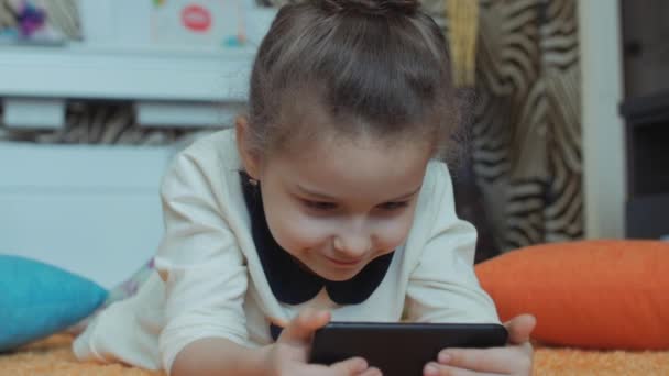 Hermosa niña jugando en el teléfono, acostada en una alfombra de punto, jugando con una tableta . — Vídeo de stock