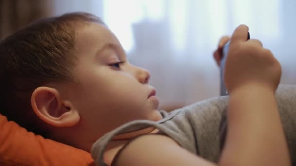 CU visage heureux de l'enfant, un petit garçon est couché sur le canapé téléphone portable, enfance heureuse . — Video