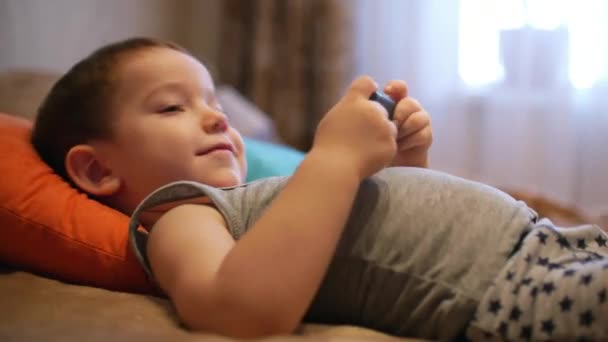 Cu blij gezicht van het kind, een kleine jongen is liggend op de Bank mobiele telefoon, gelukkige jeugd. — Stockvideo
