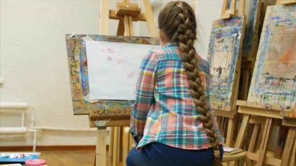 Joven artista femenina linda está en un estudio de arte, sentado detrás de un caballete y pintura sobre lienzo. Proceso de dibujo: en el estudio de arte de los artistas Hand Art Girl con un pincel Pintura sobre lienzo . — Vídeo de stock