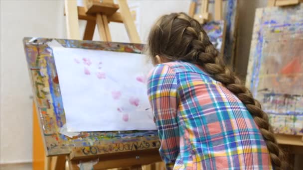 Jovem artista feminina bonito está em um estúdio de arte, sentado atrás de um cavalete e pintura na tela. Processo de Desenho: no Estúdio de Arte dos Artistas Mão Arte Menina com Pincel Pintura sobre Tela . — Vídeo de Stock