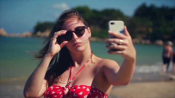 Joven hermosa morena de pie en la playa en verano, mira su teléfono y se hace selfies, en el fondo cielo azul y el mar azul, en Vietnam a la luz del día brillante . — Vídeos de Stock