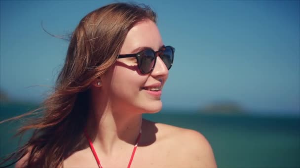 En una playa tropical Primer plano Retrato de europea hermosa linda morena con gafas de sol mujer joven o alegre chica mirando en la cámara, soplando viento pelo el viento, cámara lenta . — Vídeos de Stock