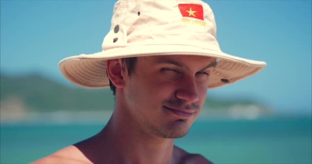 Close-up portret jonge Kaukasische van knappe brunette man in Hat, op zoek in de camera attent, glimlacht pensive, op blauwe hemel achtergrond. — Stockvideo