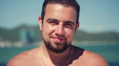 Yakın çekim portre Genç beyaz yakışıklı esmer adam bir sakal kamera bakıyor düşünceli, gülümsüyor Pensive, mavi gökyüzü arka planda.