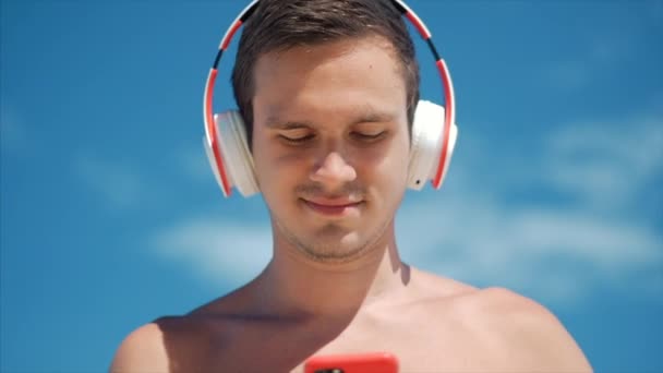 Yakışıklı genç adam kablosuz beyaz kulaklıklar onun Smartphone müzik dinleme, arka plan mavi gökyüzü plajda dans. — Stok video