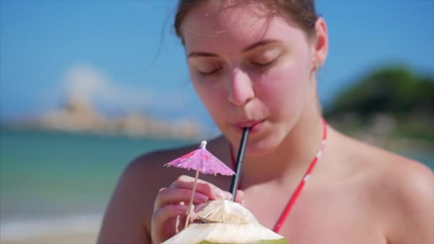 Close-up portret van een Europese mooie schattige brunette jonge vrouw of een vrolijk meisje, drink een kokosnoot op de zon, in de wind op een tropisch strand. Concept Sea Waves ontspannen door de zee in de zomer. — Stockvideo