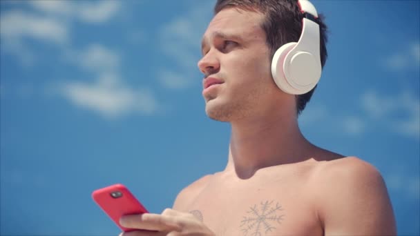 Hombre joven guapo escuchando música desde su teléfono inteligente en auriculares blancos inalámbricos, bailando en la playa en el fondo Blue Sky . — Vídeo de stock