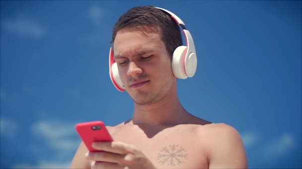 Przystojny młody człowiek słuchanie muzyki z jego smartphone w Wireless White Headphones, taniec na plaży w tle błękitne niebo. — Wideo stockowe
