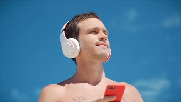 Hombre joven guapo escuchando música desde su teléfono inteligente en auriculares blancos inalámbricos, bailando en la playa en el fondo Blue Sky . — Vídeos de Stock