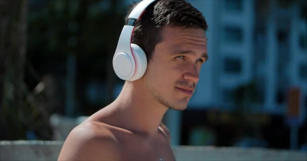 Joven guapo escuchando música desde su teléfono inteligente con auriculares blancos inalámbricos, bailando en la playa urbana en el fondo Blue Sky . — Vídeos de Stock