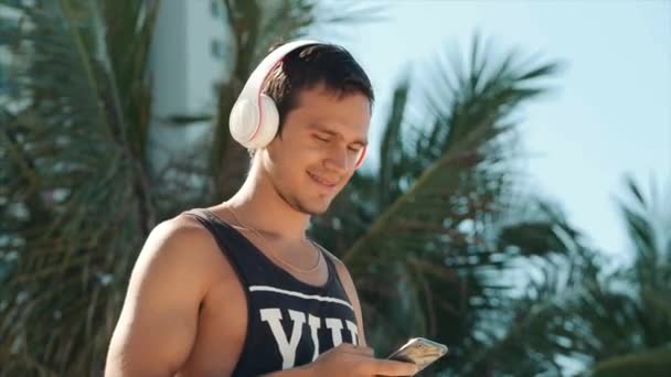 Joven guapo escuchando música desde su teléfono inteligente con auriculares blancos inalámbricos, bailando en la playa urbana en el fondo Blue Sky . — Vídeos de Stock