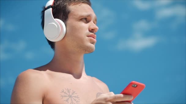 Knappe jonge man luisteren muziek van zijn smartphone in draadloze witte koptelefoon, dansen op het strand op de achtergrond Blue Sky. — Stockvideo