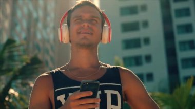 Yakışıklı genç adam kablosuz beyaz kulaklıklar onun Smartphone müzik dinleme, arka plan mavi gökyüzü Beach Urban dans.