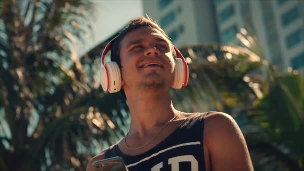 Csinos fiatalember hallgatva zene az ő smartphone vezeték nélküli fehér fejhallgató, tánc a strandon Urban a background Blue Sky. — Stock videók
