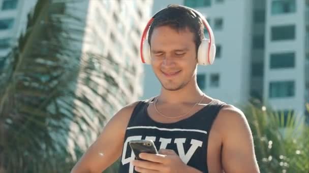 英俊的年轻人听音乐从他的智能手机在无线白色耳机, 在海滩城市在背景蓝天跳舞. — 图库视频影像