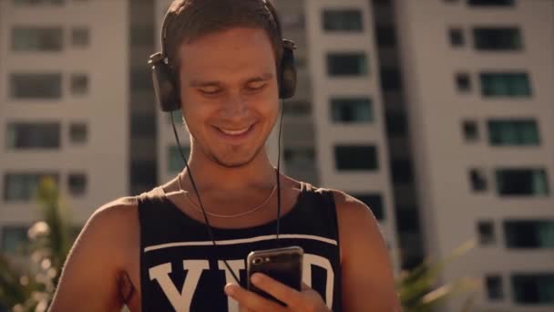 Beau jeune homme écoutant de la musique depuis son smartphone dans un casque noir sans fil, dansant sur la plage urbaine en arrière-plan Blue Sky . — Video