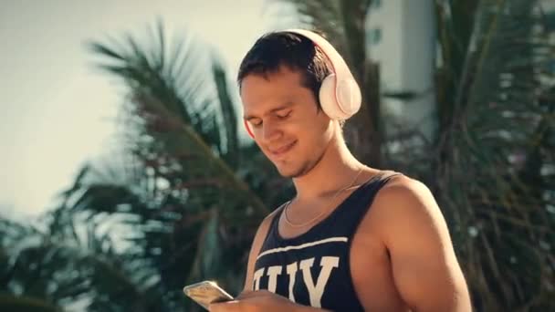 Joven guapo escuchando música desde su teléfono inteligente con auriculares blancos inalámbricos, bailando en la playa urbana en el fondo Blue Sky . — Vídeos de Stock