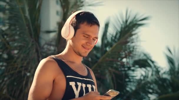 Joven guapo escuchando música desde su teléfono inteligente con auriculares blancos inalámbricos, bailando en la playa urbana en el fondo Blue Sky . — Vídeos de Stock