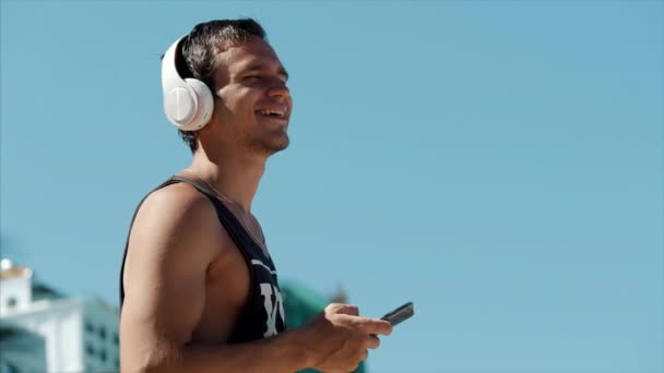 잘생긴 젊은 남자 무선 화이트 헤드폰에서 그의 스마트 폰에서 음악을 듣고, 배경 푸른 하늘에서 해변 도시에 춤. — 비디오