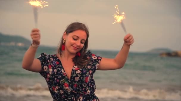 Jeune femme souriante heureuse, dansant avec scintillement au coucher du soleil au ralenti, avec feux d'artifice au coucher du soleil sur la plage . — Video