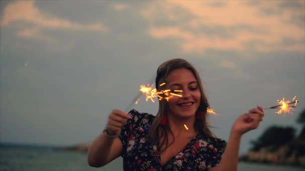 Joven mujer sonriente feliz, bailando con chispeante al atardecer en cámara lenta, con fuegos artificiales al atardecer en la playa . — Vídeos de Stock