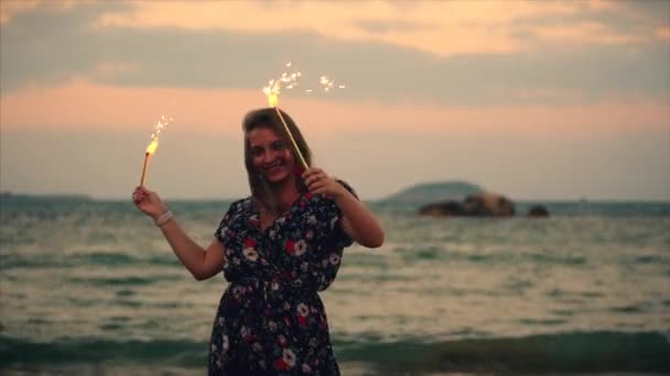 Joven mujer sonriente feliz, bailando con chispeante al atardecer en cámara lenta, con fuegos artificiales al atardecer en la playa . — Vídeos de Stock