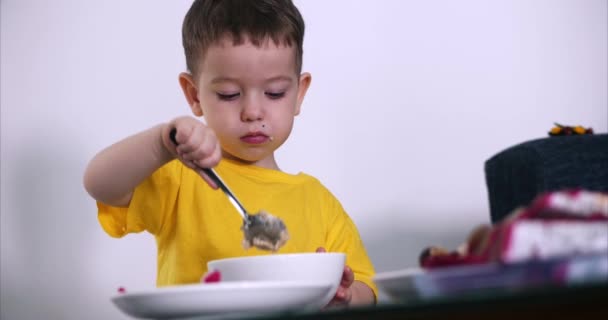 Criança pequena bonito está sentado em uma mesa e comer sua própria aveia, o bebê come de bom grado. Conceito Infância feliz . — Vídeo de Stock